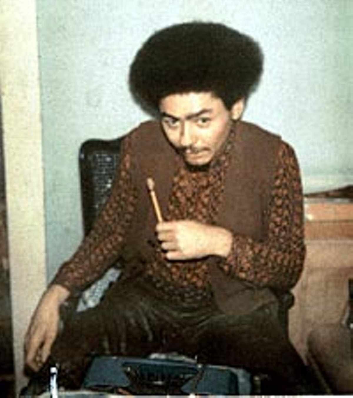 Person with an afro hairstyle sitting on a chair, wearing a patterned shirt and holding a pencil.